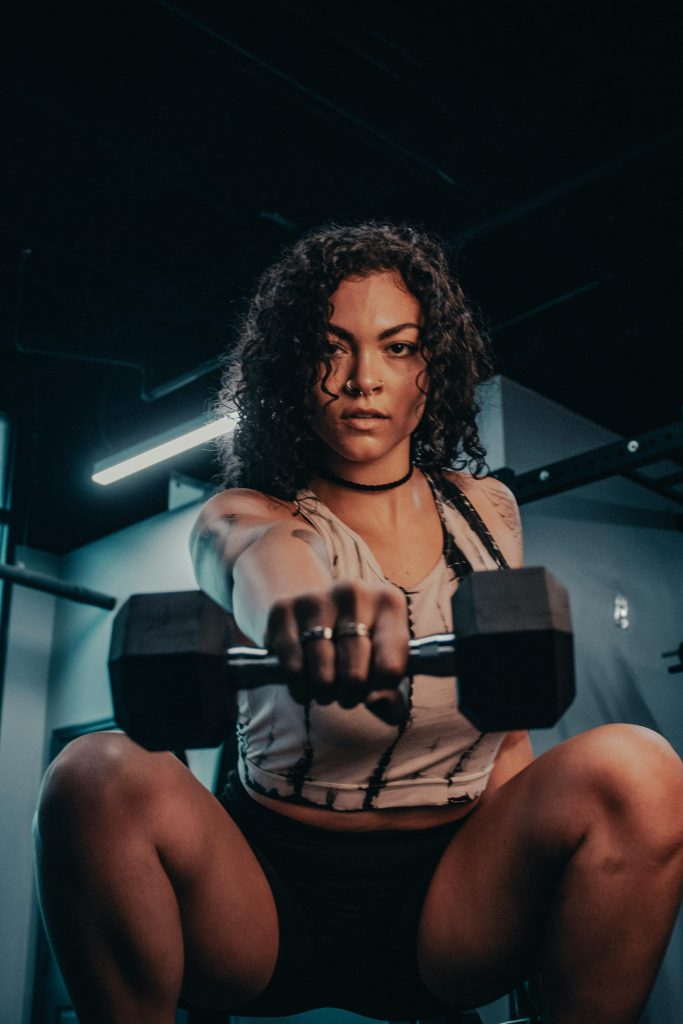pexels-photo-13197535-13197535 Focused woman with curly hair lifting a dumbbell in a dimly lit gym, showcasing strength.